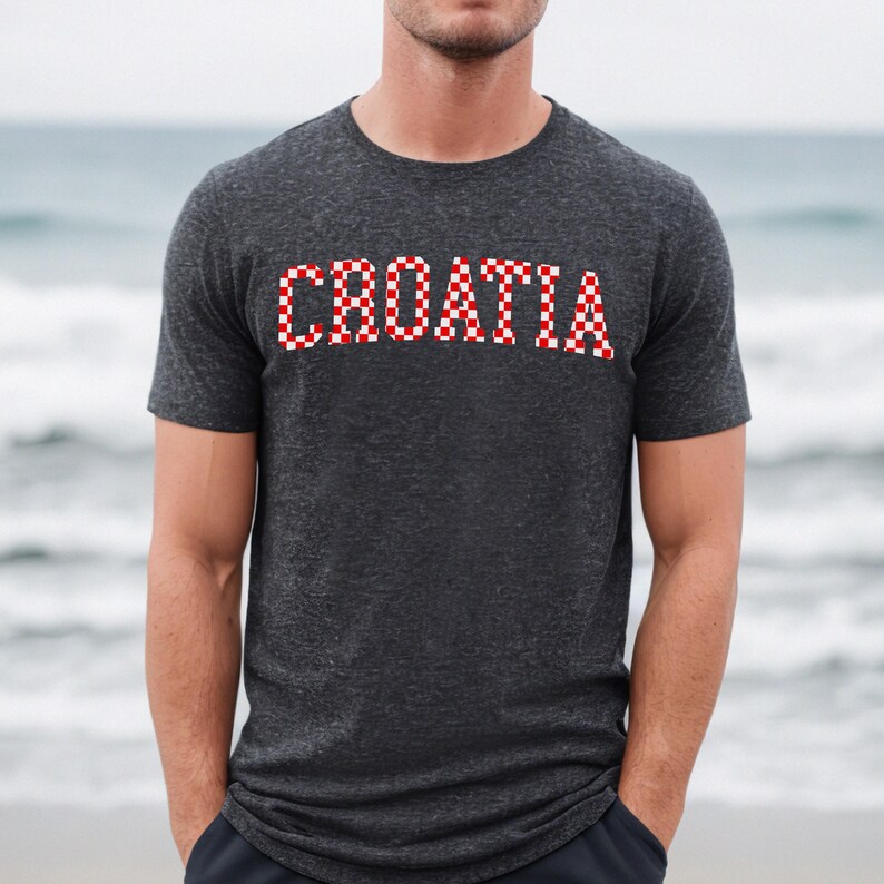 May include: Dark gray t-shirt with the word "CROATIA" in red and white checkered letters. The shirt is short-sleeved and has a crew neck. The background is a blurred beach scene.
