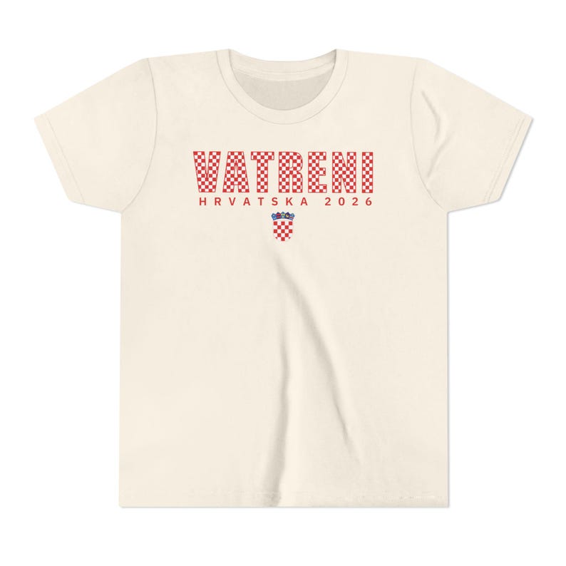May include: Cream-colored t-shirt with the word "VATRENI" in red and white checkered letters. Below, the text "HRVATSKA 2026" is printed in red. A Croatian coat of arms is centered below the text.