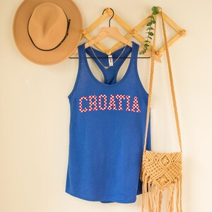 May include: A blue tank top with the word "CROATIA" in red and white checkered letters. The tank top is hanging on a wooden hanger. A tan hat and a woven purse are also visible.