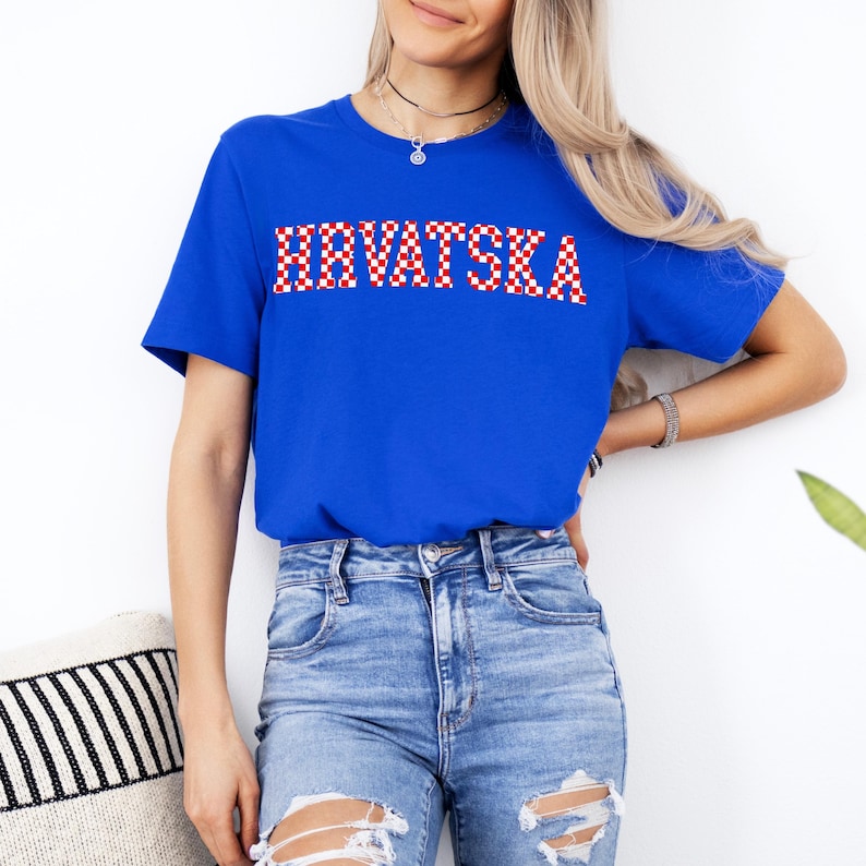Pode incluir: Uma camiseta azul royal com a palavra "HRVATSKA" em um padr&atilde;o xadrez vermelho e branco no peito. A camisa &eacute; um estilo de gola redonda de manga curta. A modelo est&aacute; usando jeans azuis.