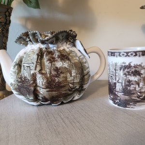 May include: A white teapot with a brown and green floral pattern, and a white mug with a similar pattern. The teapot has a ruffled fabric cover.