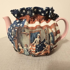 May include: A teapot cozy with a red, white, and blue American flag design. The cozy features a scene of women sewing an American flag.