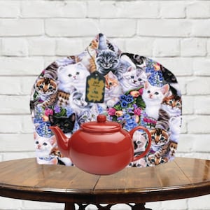 May include: A red teapot with a cat-themed tea cozy. The cozy is covered in a pattern of colorful cats, flowers, and the text "Tea Time Meow".