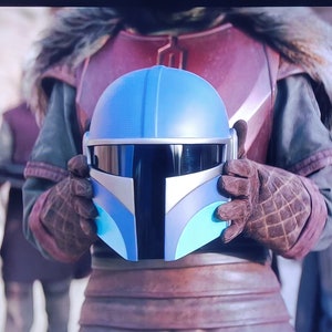 May include: A blue and white Mandalorian helmet with a black visor. The helmet is held by a person wearing brown gloves and a red and brown outfit. The helmet has a light blue dome and a dark blue and white faceplate.