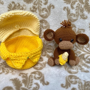 May include: A crocheted monkey toy holding a crocheted banana. The monkey is brown with black eyes and a yellow banana. The monkey is sitting next to a yellow crocheted basket with a lid.