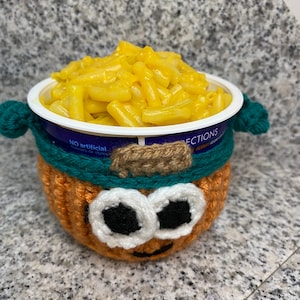 May include: A crocheted orange pumpkin-shaped cup with a green and white hat and black eyes holds a container of macaroni and cheese. The container has the text "NO artificial flavours or dyes" and "LECTIONS" on it.