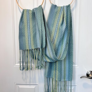 May include: Two blue and green woven scarves with fringe hanging from wooden hoops. The scarves feature vertical stripes in shades of blue and green. The fringe at the bottom of the scarves is a mix of blue and green.
