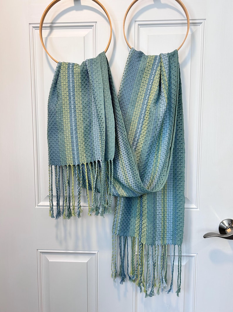 May include: Two blue and green woven scarves with fringe hanging from wooden hoops. The scarves feature vertical stripes in shades of blue and green. The fringe at the bottom of the scarves is a mix of blue and green.