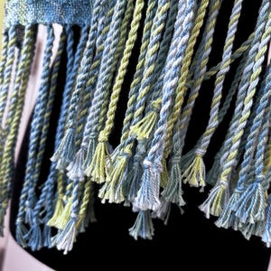 May include: Close-up of a woven textile with blue, green, and white twisted fringe. The fringe is knotted at the top and has small tassels at the bottom. The textile appears to be a scarf or shawl.
