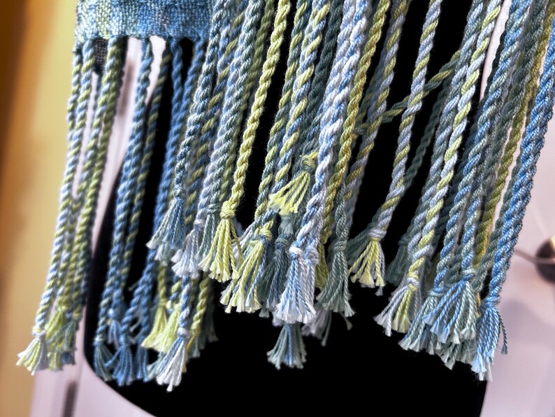 May include: Close-up of a woven textile with blue, green, and white twisted fringe. The fringe is knotted at the top and has small tassels at the bottom. The textile appears to be a scarf or shawl.