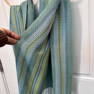 May include: A handwoven scarf in shades of blue, green, and yellow hangs from a wooden hoop. The scarf has a textured pattern with vertical stripes and fringe at the bottom. The colour palette is soft and muted, creating a calming aesthetic.