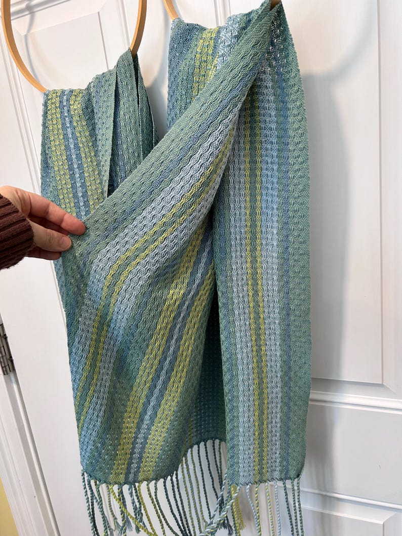 May include: A handwoven scarf in shades of blue, green, and yellow hangs from a wooden hoop. The scarf has a textured pattern with vertical stripes and fringe at the bottom. The colour palette is soft and muted, creating a calming aesthetic.