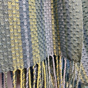 May include: Close-up of a woven textile with a textured pattern in shades of blue, green, and yellow. The fabric has a fringed edge. The weave creates a geometric design.