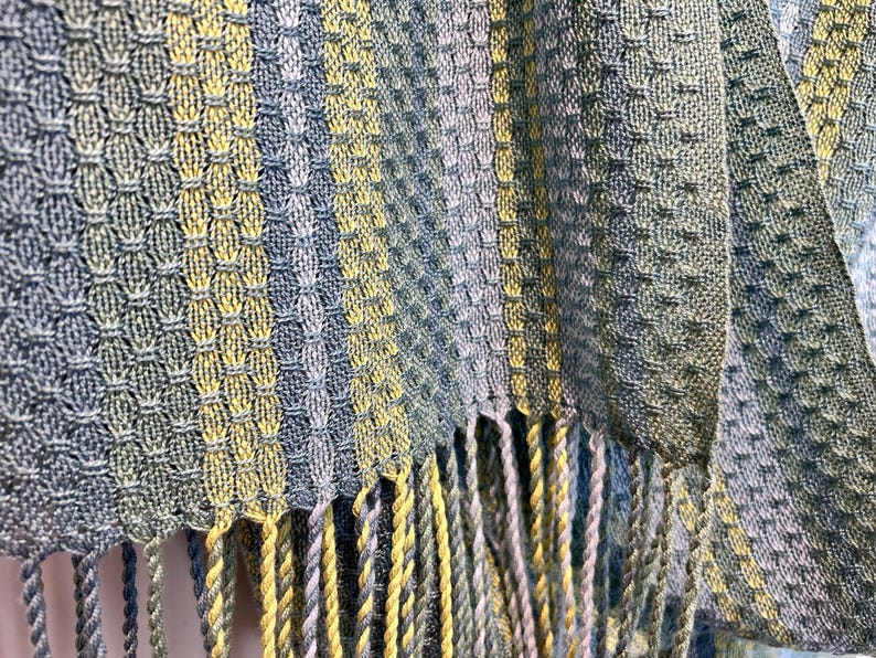 May include: Close-up of a woven textile with a textured pattern in shades of blue, green, and yellow. The fabric has a fringed edge. The weave creates a geometric design.