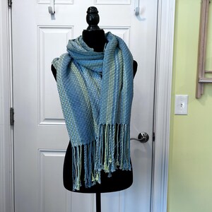 May include: A blue and green woven scarf, draped on a black mannequin. The scarf features a textured design and fringed edges. The colour scheme includes light and dark blues with touches of green.