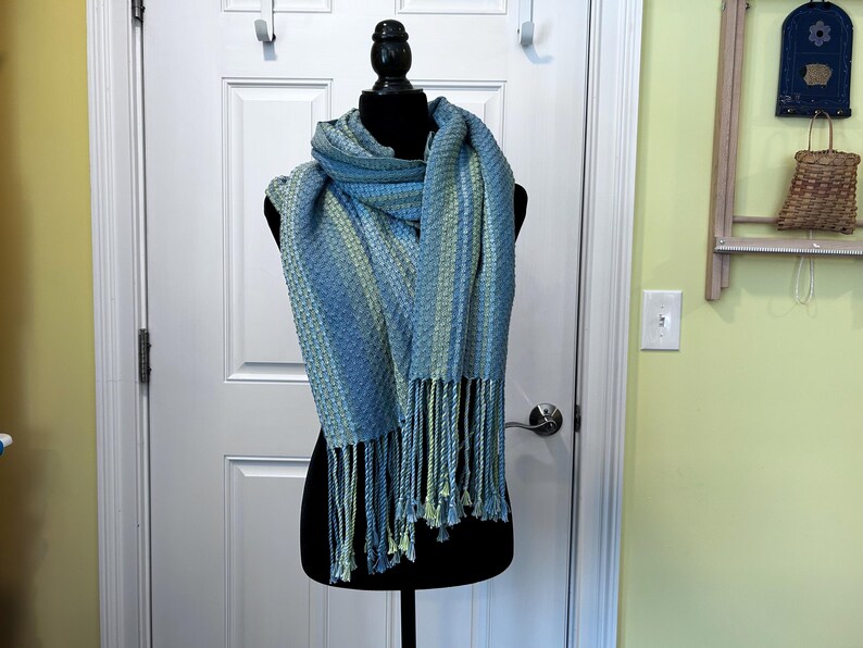 May include: A blue and green woven scarf, draped on a black mannequin. The scarf features a textured design and fringed edges. The colour scheme includes light and dark blues with touches of green.