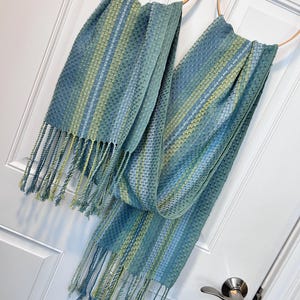 May include: Two blue and green woven scarves with fringe hanging from wooden hoops. The scarves have a striped pattern with varying shades of blue and green. The fringe is a mix of blue and yellow-green.