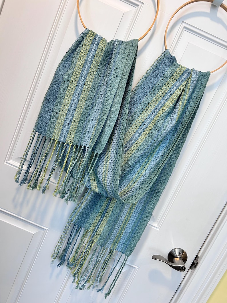 May include: Two blue and green woven scarves with fringe hanging from wooden hoops. The scarves have a striped pattern with varying shades of blue and green. The fringe is a mix of blue and yellow-green.