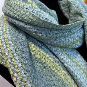May include: A close-up of a woven scarf in shades of blue, green, and yellow. The scarf has a textured pattern with alternating stripes of colour. The fabric appears soft and drapes gently.