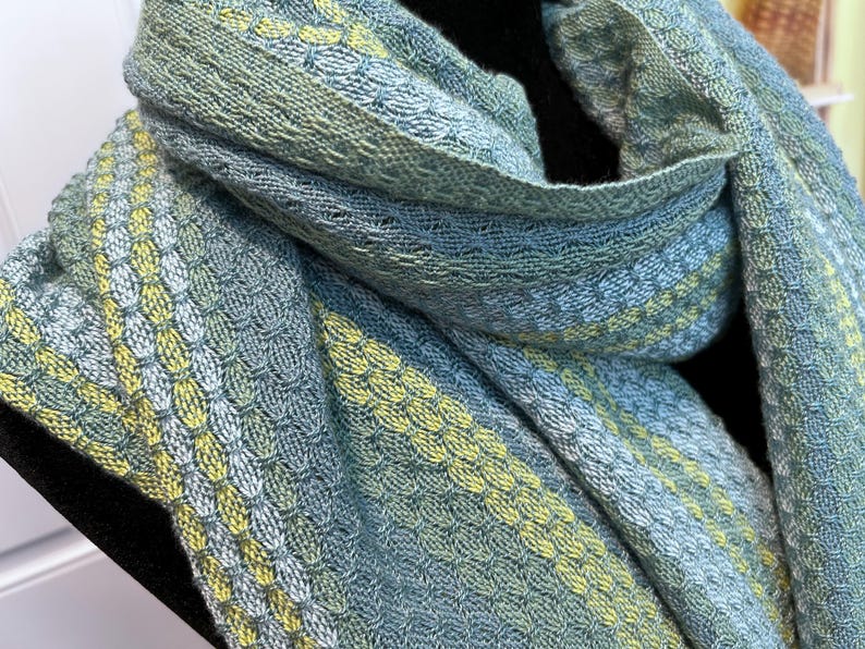 May include: A close-up of a woven scarf in shades of blue, green, and yellow. The scarf has a textured pattern with alternating stripes of colour. The fabric appears soft and drapes gently.