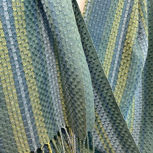 May include: A woven scarf in teal, green, and yellow hues. The scarf has a textured pattern with vertical stripes and a fringed edge. The fabric appears soft and drapes gently, suitable for wear.