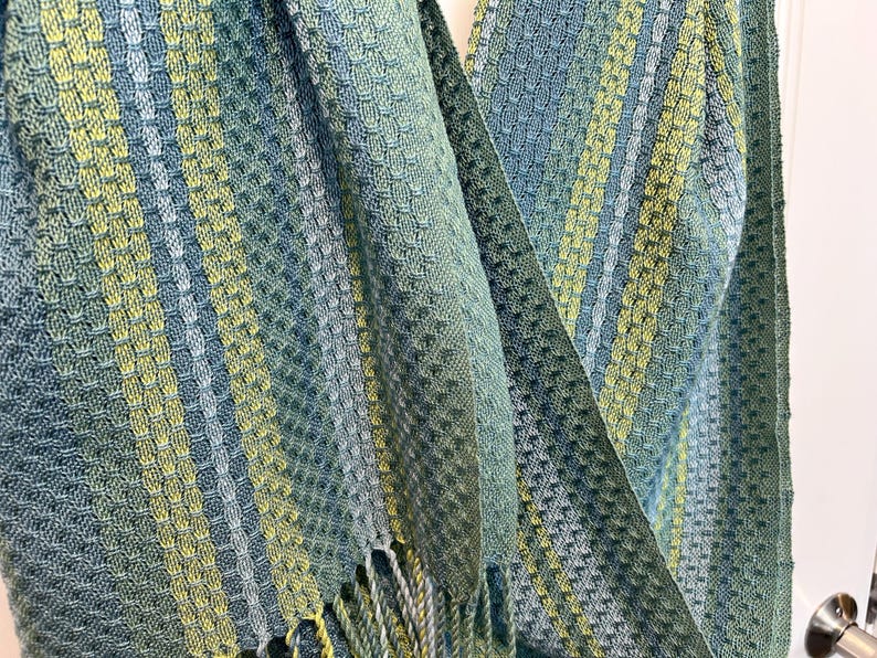 May include: A woven scarf in teal, green, and yellow hues. The scarf has a textured pattern with vertical stripes and a fringed edge. The fabric appears soft and drapes gently, suitable for wear.