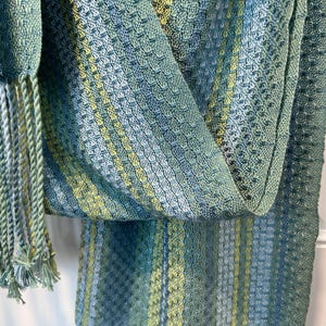 May include: A close-up shot of a woven scarf with a textured pattern. The scarf displays shades of blue, green, and yellow, with a fringe of twisted yarn in matching colours. The fabric appears soft and lightweight, suitable for a shawl or wrap.