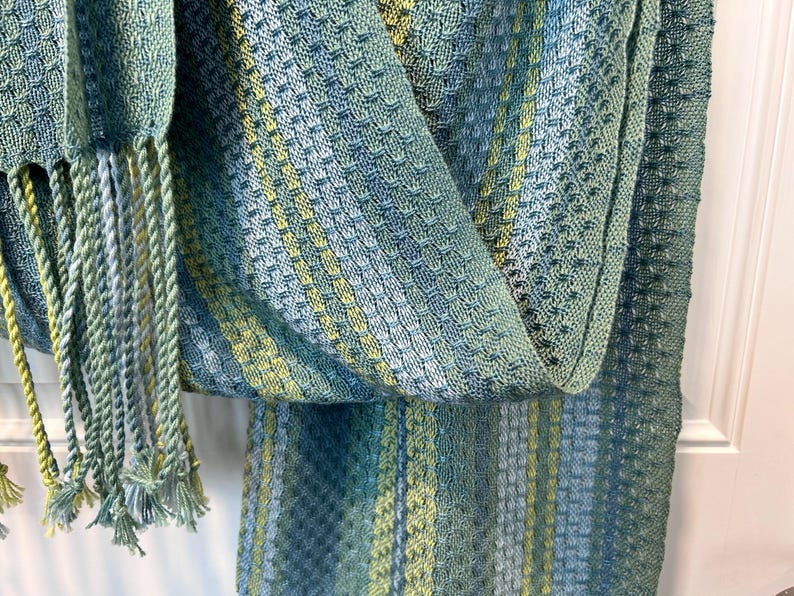 May include: A close-up shot of a woven scarf with a textured pattern. The scarf displays shades of blue, green, and yellow, with a fringe of twisted yarn in matching colours. The fabric appears soft and lightweight, suitable for a shawl or wrap.