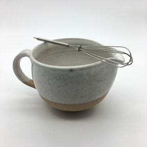 May include: A white ceramic mixing bowl with a speckled glaze and a brown rim. The bowl has a handle and a spout. A stainless steel whisk is resting on the rim of the bowl.