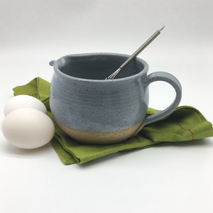 May include: A gray ceramic mixing bowl with a handle and a speckled glaze. The bowl is sitting on a green napkin with two white eggs beside it. A metal whisk is inside the bowl.