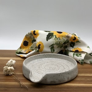 May include: White ceramic spoon rest with a speckled finish. The rest has a curved shape with a spout for draining excess liquid. The spoon rest is sitting on a wooden surface.
