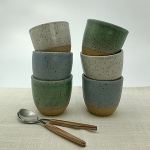 May include: A stack of six speckled ceramic mugs in three colors: green, blue, and white. The mugs are stacked in pairs, with the top mug of each pair being a different color. The mugs are all speckled with brown and white flecks.