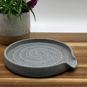 May include: A gray ceramic spoon rest with a speckled finish and a swirl design. The rest has a small spout on the side.