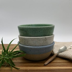 May include: Three ceramic bowls stacked on top of each other. The top bowl is green with white speckles, the middle bowl is white with black speckles, and the bottom bowl is blue with white speckles.