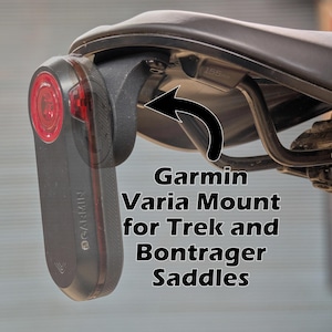 May include: A black Garmin Varia mount attached to a bicycle seat. The mount is designed for Trek and Bontrager saddles. The Garmin device has a red light and is attached to the mount.