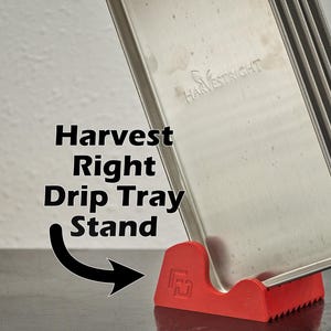 May include: A stack of stainless steel Harvest Right drip trays, supported by a red plastic stand. The stand has a unique design with a textured base and the text "Harvest Right Drip Tray Stand" is visible.