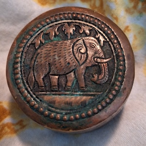 Op de afbeelding: Een ronde koperen container met een in reliëf uitgesneden olifant in het midden. Het deksel is versierd met een kralenrand en gestileerde bladmotieven boven de olifant. Het metalen oppervlak vertoont een verweerde patina met groene oxidatie.