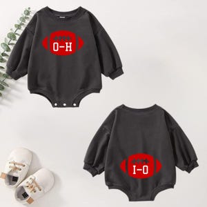 May include: Two dark grey baby bodysuits with long sleeves. Each bodysuit features a red football graphic with white stitching and the text "O-H" and "I-O". The bodysuits are displayed on a light background with a pair of white baby shoes.