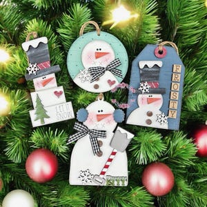 May include: A collection of wooden snowman ornaments with various designs. Each ornament features a snowman with a carrot nose, black eyes, and a checkered bow. Some ornaments have the text "LET IT SNOW" or "FROSTY".