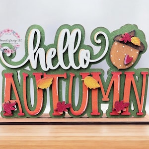 May include: A wooden sign with the words "hello autumn" in white letters on a green background. The sign is decorated with fall leaves and a small acorn with a red heart.