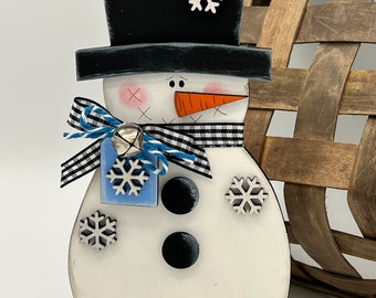 May include: A wooden snowman figurine with a black top hat, a carrot nose, and black eyes. The snowman is decorated with white snowflakes and a black and white checkered scarf. The snowman is standing on a black base.