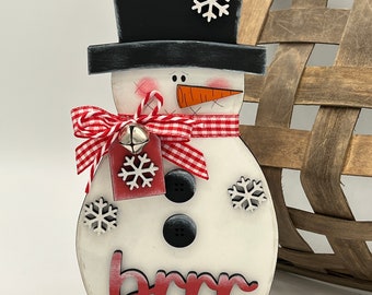 May include: A white wooden snowman with a black top hat, red and white gingham bow, and a carrot nose. The snowman has black buttons, a snowflake tag, and the word "brrr" written in red letters.