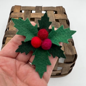 May include: A handmade Christmas holly decoration with green felt leaves and three red and pink felt berries. The holly is held in front of a brown woven basket, suggesting a holiday theme.