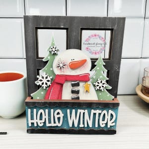 May include: A wooden winter-themed decoration featuring a snowman with a red scarf, orange carrot nose, and black coal buttons. The decoration includes green trees, snowflakes, and the text "HELLO WINTER" on a blue base.