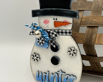 May include: A wooden snowman decoration with a black top hat, a checkered scarf, and a carrot nose. The snowman has three black buttons and snowflake accents. The word "winter" is written in blue script on the base. A small bell and blue ribbon are attached.