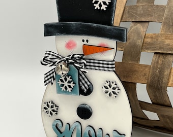 May include: A wooden snowman figurine with a black top hat, a white body, and a blue and white scarf. The snowman has a carrot nose, black eyes, and a black button. The snowman is holding a blue tag with a snowflake on it. The snowman is standing on a black base. The word "snow" is written in blue on the snowman's body.