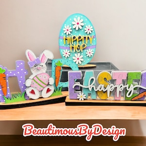 May include: Wooden Easter sign with the words "Happy Easter" in white letters. The sign is decorated with purple, yellow, green, and blue paint and has a glittery finish. There is a bunny holding a pink and green egg and a blue egg with the words "Hippity Hop" on it.
