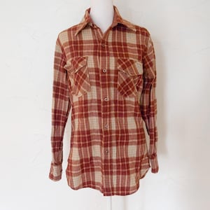 Puede incluir: Camisa de manga larga con estampado de cuadros rojos, beige y marrón. Tiene dos bolsillos en el pecho y un cuello clásico. La tela parece ser un material suave y tejido. La camisa se muestra en un maniquí.