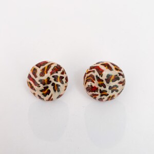 May include: Two round earrings with a cream-colored base and a pattern of red, yellow, and black shapes. The earrings are set against a white background.