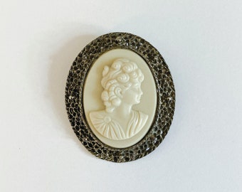May include: A white cameo brooch featuring a woman's profile with a detailed hairstyle. The cameo is set in a silver-toned metal frame with a decorative, intricate design.